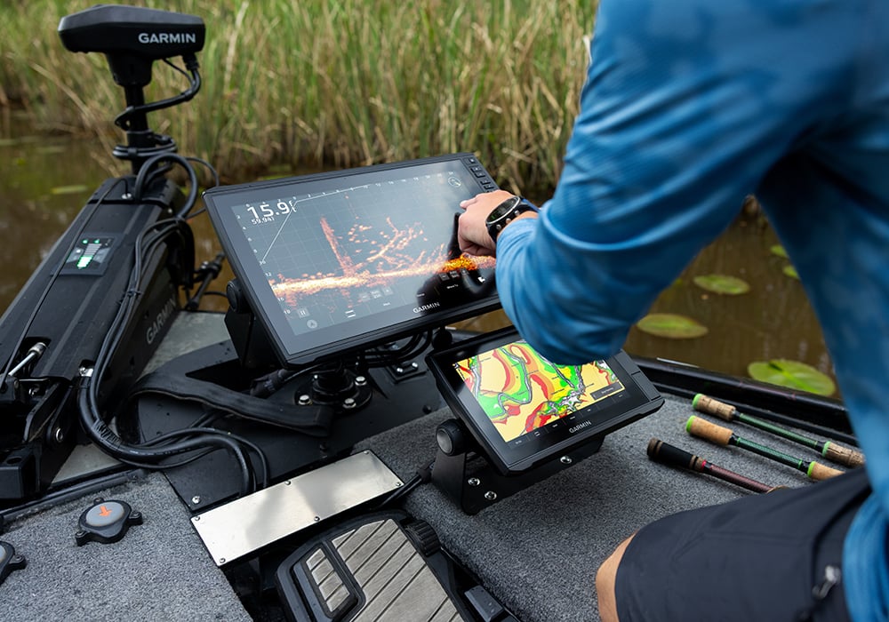 Angler on a boat kneeling to adjust the touchscreen of his Garmin chartplotter while it depicts sonar data. The boat is in the middle of a marsh with more Garmin equipment onboard.