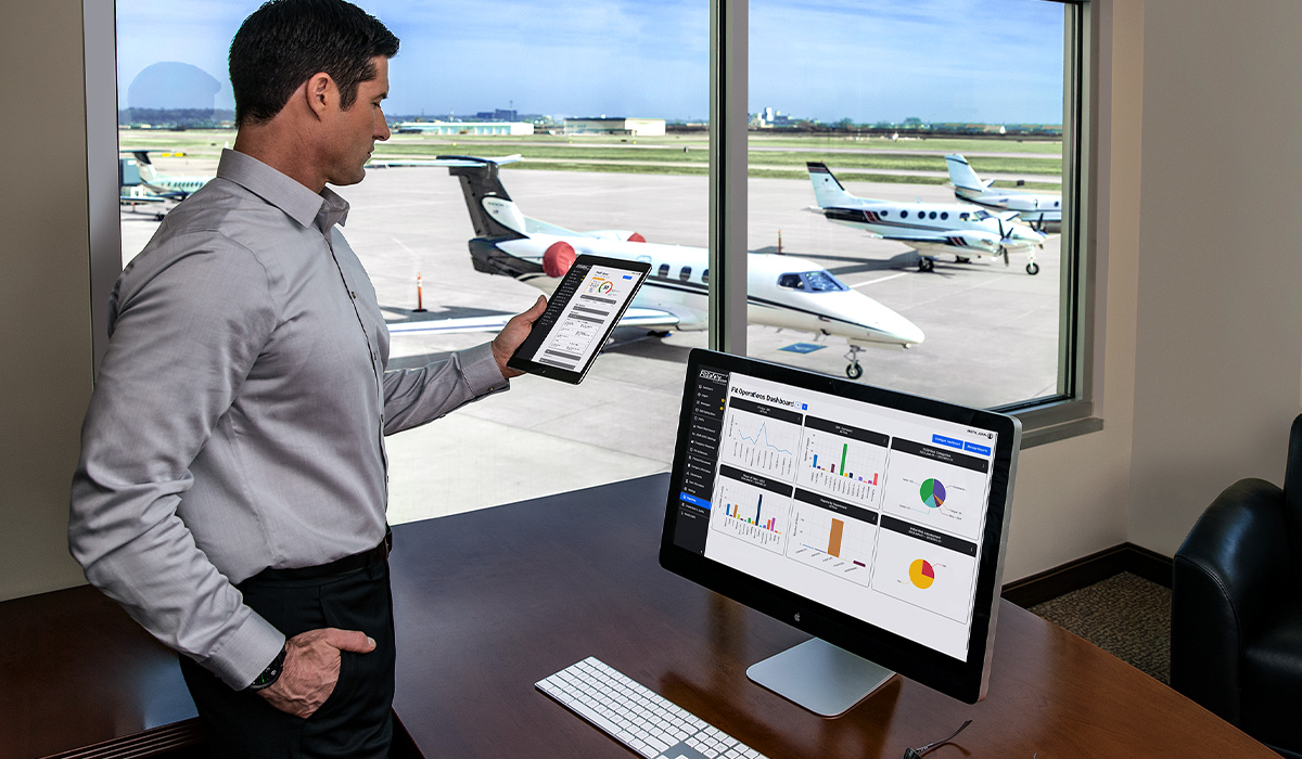 A pilot looks over his FltPlan SMS performance dashboard on his devices inside an office. In the background, airplanes are parked at a hangar.