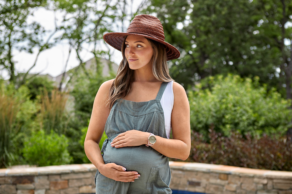 A pregnant woman wears green overalls and a brown hat, placing her hands over her belly.